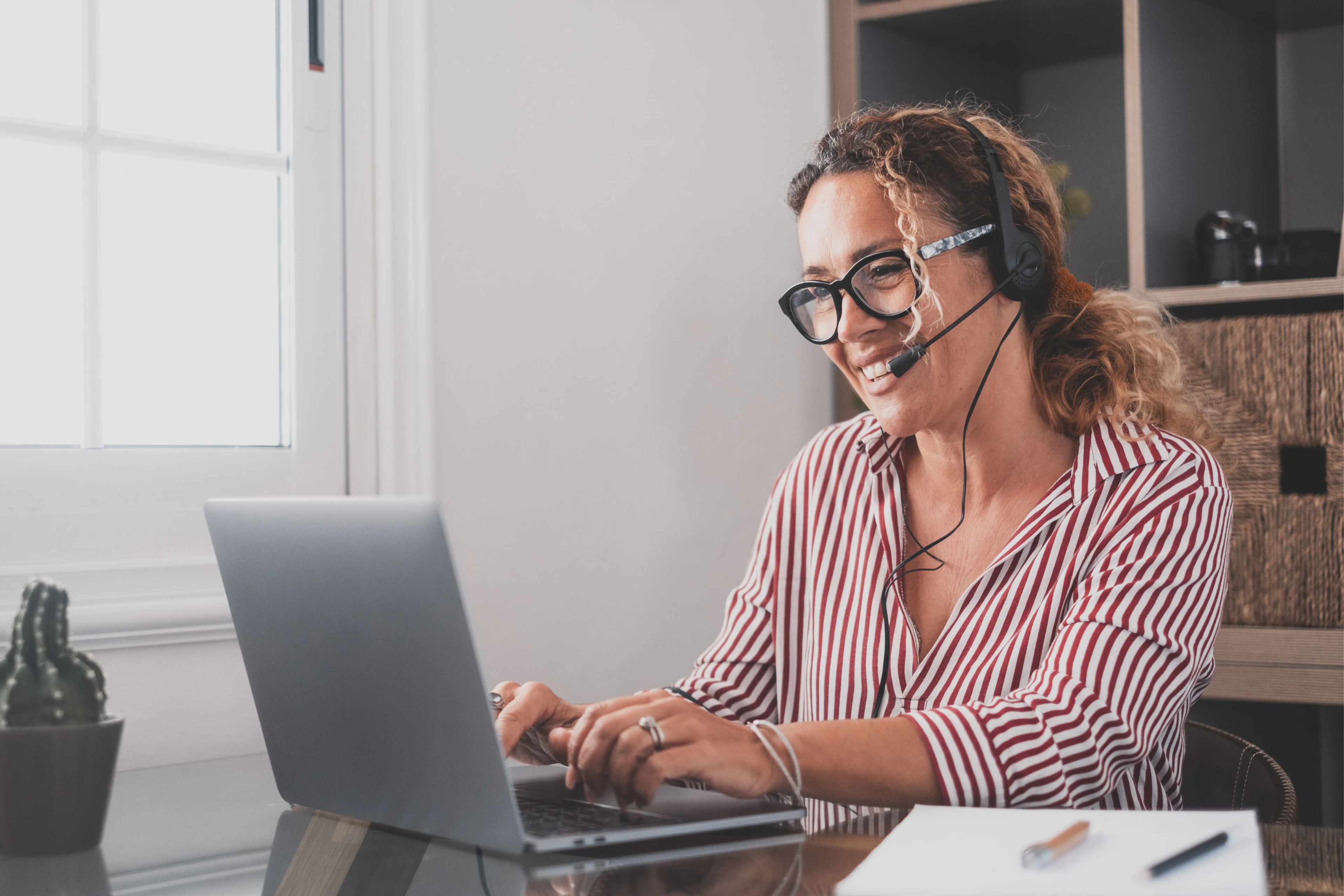 Frau mit Headset arbeitet am Laptop an einem Schreibtisch am Fenster