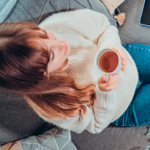 Frau entspannt mit einer Tasse Tee auf dem Sofa, während ein Laptop in der Nähe steht.